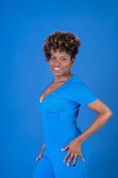 A woman in a blue dress is smiling and posing for the camera. The blue dress is a tight-fitting jumpsuit with a patterned design. The woman's smile and confident posture convey a sense of happiness