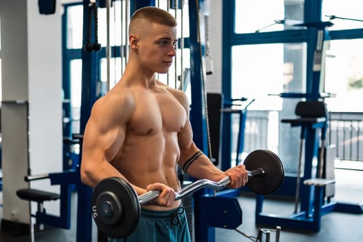A shirtless bodybuilder trains his biceps using a barbell in a gym