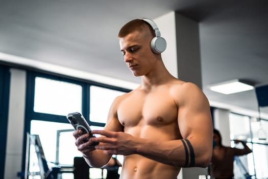 A shirtless bodybuilder in a gym, wearing headphones, looks at his phone.