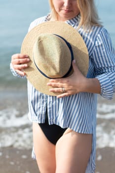 Young blonde woman in fashionable white blue striped beach shirt with straw hat posing against the background of the sea on the shore, copy space
