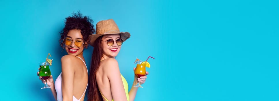 Two women in sunglasses and summer attire pose with cocktails in hand against a bright blue background. The image captures the essence of a fun and carefree summer day, copy space