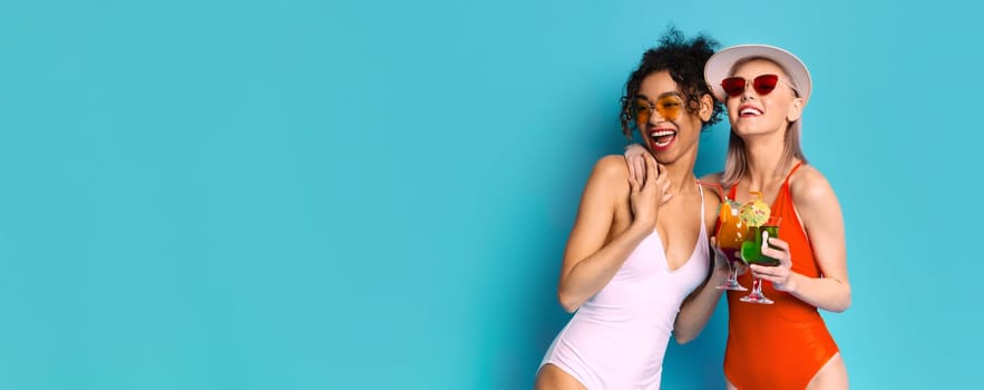 Two women in swimsuits wearing sunglasses and smiling while holding cocktails on a blue background. They are standing side-by-side and appear to be friends enjoying a fun day at the beach or pool.