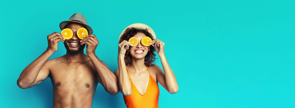 African American happy couple is posing against a vibrant blue background. They are both wearing summery attire and holding orange slices over their eyes, copy space