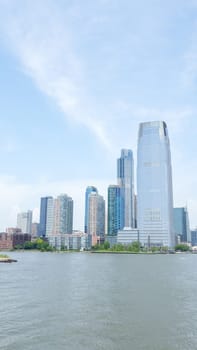 An urban park with a pedestrian walkway and green lawn overlooks a wide river with a city skyline in the background. The park has a modern minimalist design with a large concrete wall.