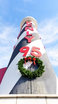 Tall black-and-white lighthouse sign with red accents in front of a historic building, under a clear blue sky with white clouds. Gas pumps and cars in parking lot by the lighthouse.