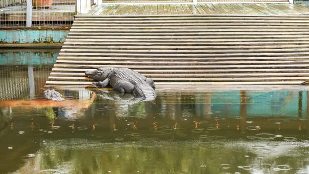 A large crocodile with a long tail and sharp teeth is lying on the shore of a lake. The crocodile is dark gray in color and has a rough, scaly skin. It is sunning itself on a warm rock.