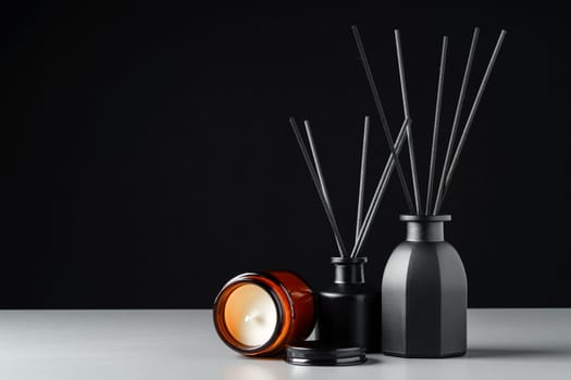 A black reed diffuser bottle with black reeds is placed on a grey surface with a candle next to it. The background is a solid black.