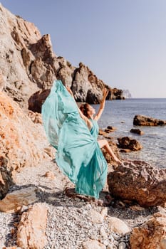 Woman green dress sea. Woman in a long mint dress posing on a beach with rocks on sunny day. Girl on the nature on blue sky background