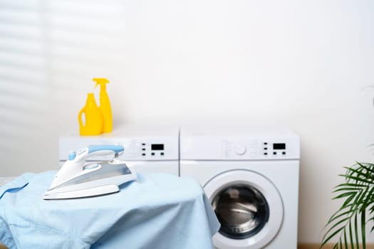A clean and modern laundry room featuring two white washing machines, an ironing board with a blue shirt draped over it, a pile of colorful clothes on top of one of the machines, and a potted houseplant in the corner.