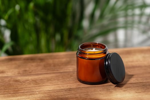 A close-up image of an amber glass candle jar on a wooden table with green foliage blurred in the background.