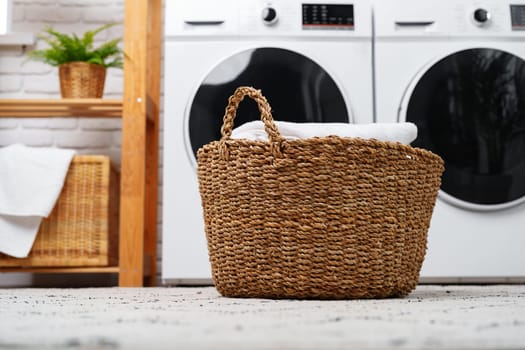 A wicker laundry basket with clean white towels sits on a light grey patterned rug near two modern white washers in a home.