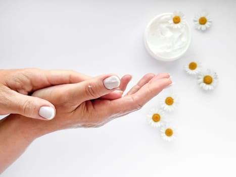 Close up of middle age woman hands applying cream with chamomile flowers and hand cream container on white background. Skincare and natural beauty concept. For healthcare, wellness, organic product.