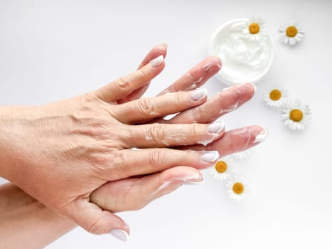 Close up of middle age woman hands applying cream with chamomile flowers and hand cream container on white background. Skincare and natural beauty concept. For healthcare, wellness, organic product.