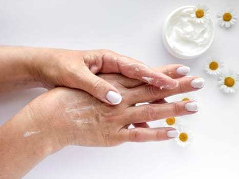 Close up of middle age woman hands applying cream with chamomile flowers and hand cream container on white background. Skincare and natural beauty concept. For healthcare, wellness, organic product.