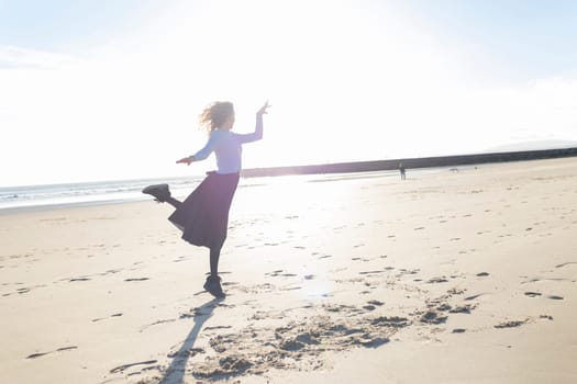 A woman is running on the beach with her arms outstretched. The sun is shining brightly, creating a warm and inviting atmosphere. The beach is empty