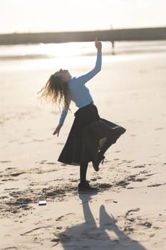 A woman is dancing on the beach with her arms outstretched. The beach is empty and the sun is shining brightly