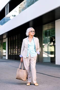 stylish mature woman with a bag in her hand standing next to a building, concept of urban lifestyle in adulthood, copyspace for text