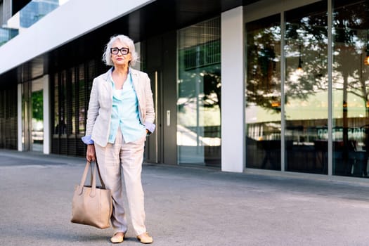 stylish mature woman with a bag in her hand standing next to a building, concept of urban lifestyle in adulthood, copyspace for text