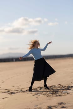 A woman is standing on the beach, wearing a black skirt and a blue shirt. She is dancing and she is enjoying herself. The beach setting and the woman's carefree attitude create a sense of relaxation
