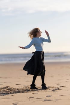 A woman is standing on the beach, wearing a blue shirt and black skirt. She is dancing and she is enjoying herself. The beach setting and the woman's carefree attitude create a sense of relaxation