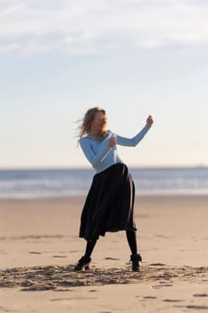 A woman is dancing on the beach in a black skirt. She is wearing a blue shirt and black boots