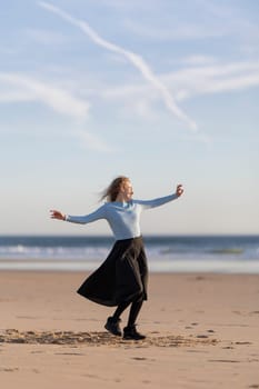 A woman is standing on the beach, wearing a blue shirt and black skirt. She is smiling and she is enjoying herself. The beach is a beautiful and peaceful setting, with the ocean in the background