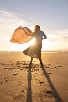 A woman stands on a beach with a scarf blowing in the wind. The scene is serene and peaceful, with the sun setting in the background
