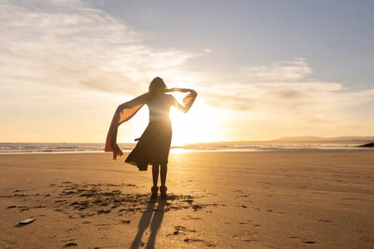 A woman stands on a beach with a scarf around her neck. The sky is a beautiful orange color, and the sun is setting. The woman is enjoying the moment and the beautiful scenery