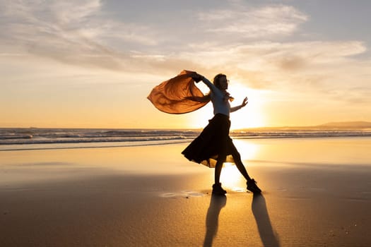 A woman is dancing on the beach with a scarf in her hand. The sky is orange and the sun is setting