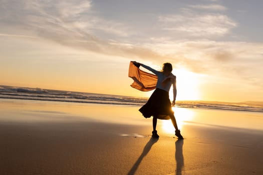 A woman is standing on the beach with a scarf around her neck. The sky is orange and the sun is setting