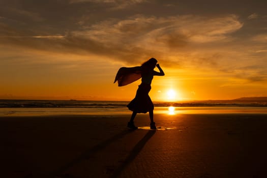 A woman is walking on the beach with a scarf in her hand. The sky is orange and the sun is setting