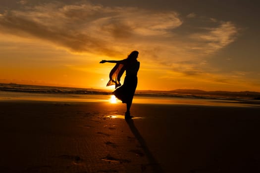 A woman is standing on the beach, with the sun setting in the background. She is wearing a scarf and she is dancing. Scene is peaceful and serene, as the woman is enjoying the beauty of the sunset