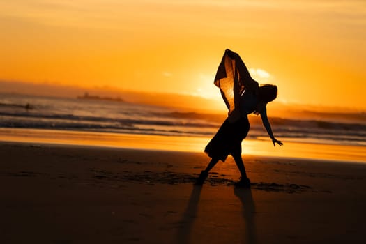A woman is dancing on the beach in the evening. She is wearing a long skirt and holding a scarf. The scene is serene and peaceful, with the sun setting in the background