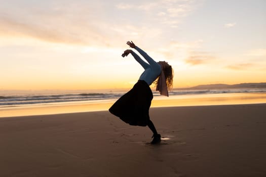 A woman is dancing on the beach in a skirt. The sky is orange and the sun is setting