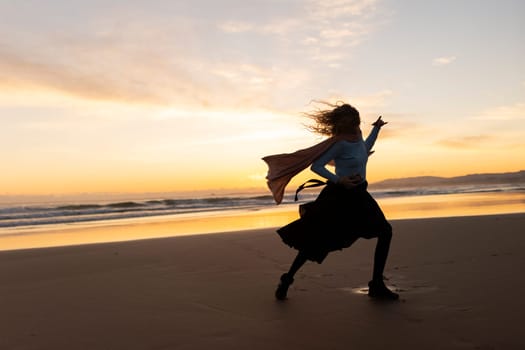 A woman is running on the beach with a scarf around her neck. The sky is orange and the water is calm