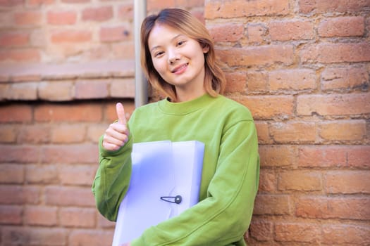 Happy smiling young man holding a notebook and pointing thumb up looking at camera at the university campus