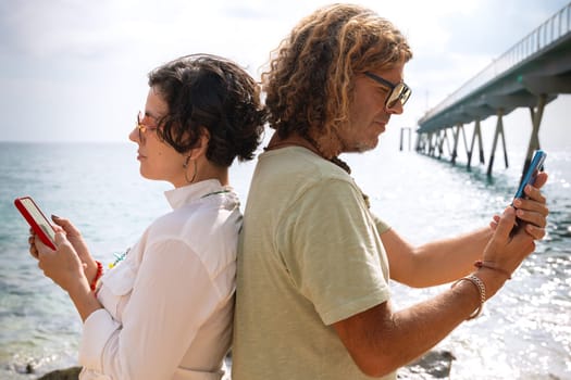 Middle-aged man and woman sitting on the beach with surprised face, browsing telephones apps. Concept:Holidays y Technology