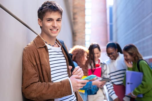 Young smiling student with notebook in hand uses social networks with smartphone applications and wireless technology looking at camera outdoors.