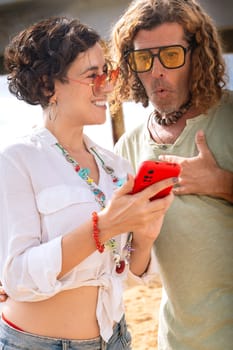 Middle-aged man and woman sitting on the beach with surprised face, browsing smartphone apps. Concept:Holidays y Technology