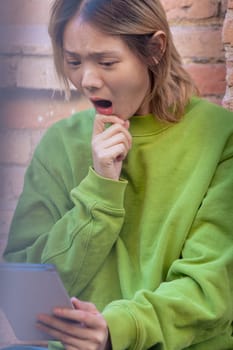 Young Chinese female student with surprised face watching social media content with digital tablet apps and wireless technology outdoors.Vertical