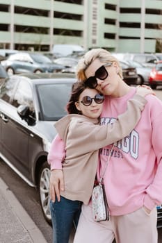 Tender moment between a mother and her daughter in an urban setting. Both are dressed in casual, fashionable outfits and wearing sunglasses, exuding a relaxed and stylish vibe.