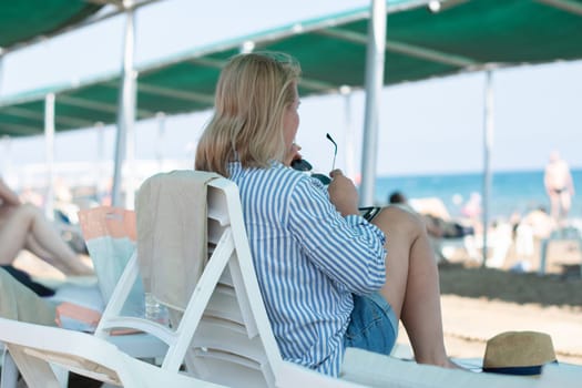 Young woman sitting on a sun lounger on the beach, putting on sunglasses and looking dreamily at the sea,woman looks thoughtful,looks into the distance,deep in thoughts,midlife crisis