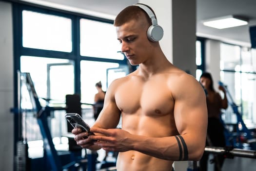 A shirtless man in a gym, wearing headphones, looks at his phone