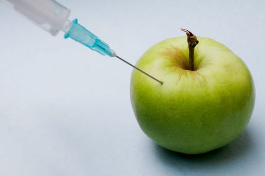 A syringe with a needle pierced a green apple close-up on a blue background. GMO concept photo.