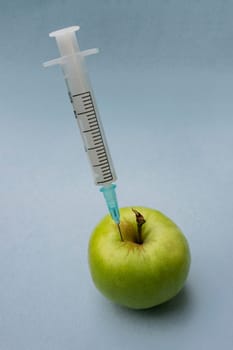 A close-up vertical photo of a syringe stuck into a green apple on a blue background. Syringe with liquid in fruit. GMO concept photo.
