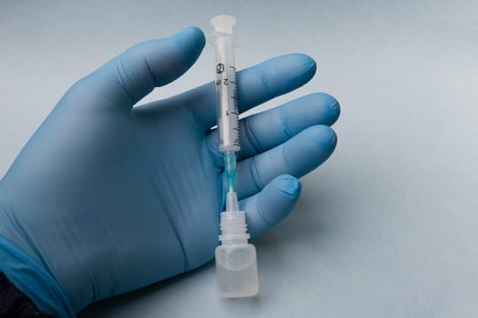 A man's hand in a blue glove holds a syringe stuck into a plastic bottle.