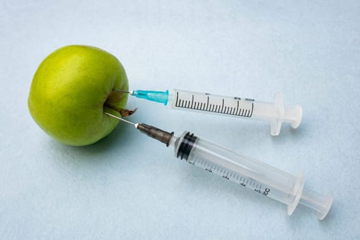Green apple with two stuck syringes on a blue background close-up. Food research concept photo. Poison enters the green apple.