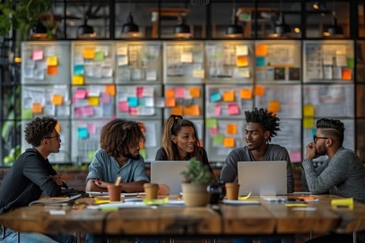 A group of diverse individuals, including a woman and three men, are collaborating on laptops during a meeting in a modern office setting. They are sitting around a large wooden table, with a wall covered in colorful sticky notes behind them. The team members are engaged in conversation, discussing ideas and working on a project together.