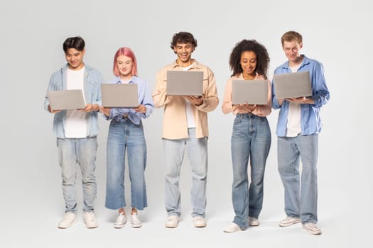 A diverse group of five young adults stand in a row, each using a laptop computer. They are all dressed casually and appear to be focused on their devices. The background is simple white