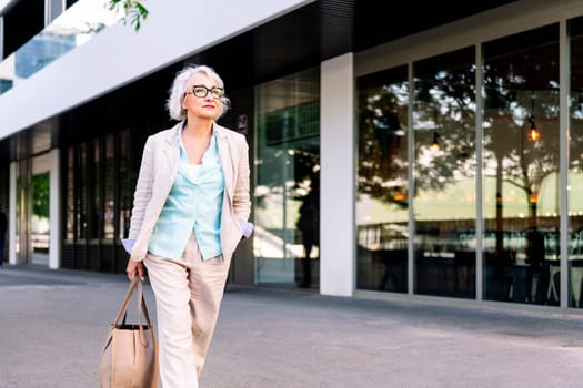 stylish mature woman walking with a bag in her hand next to a building, concept of urban lifestyle in adulthood, copyspace for text